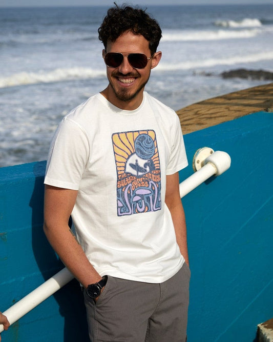 A man in sunglasses leaning on a railing by the sea, wearing a Saltrock Surf Fest Mens Short Sleeve T-Shirt in White with a distressed surf print, smiling at the camera.