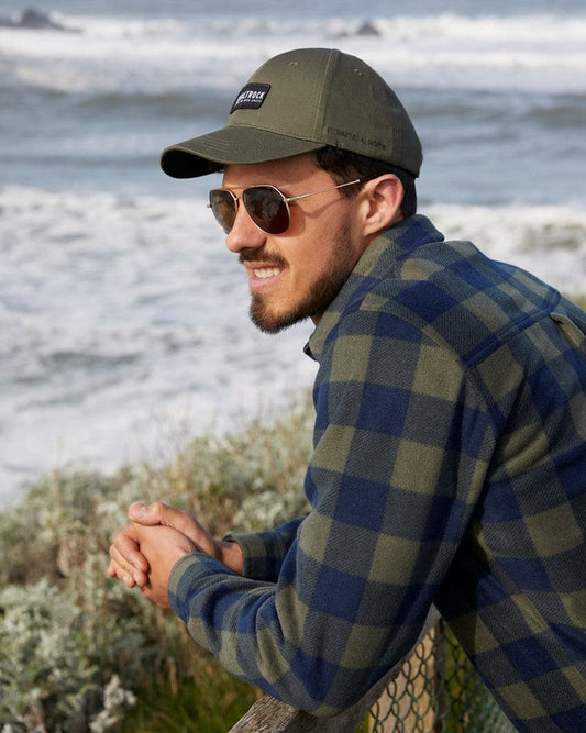Man in a Waldron - Mens Fleece Shirt in Dark Green and Saltrock cap leaning on a fence by the ocean, wearing sunglasses, smiling slightly.