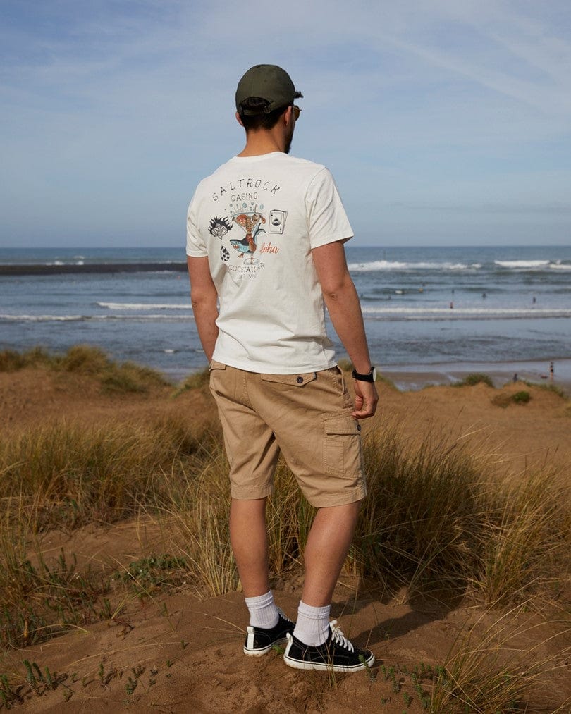 Man wearing Vegas Cocktail - Mens Short Sleeve T-Shirt - White with Saltrock branding, machine washable shorts, and a cap, standing on sand dunes overlooking a beach with surfers in the sea.