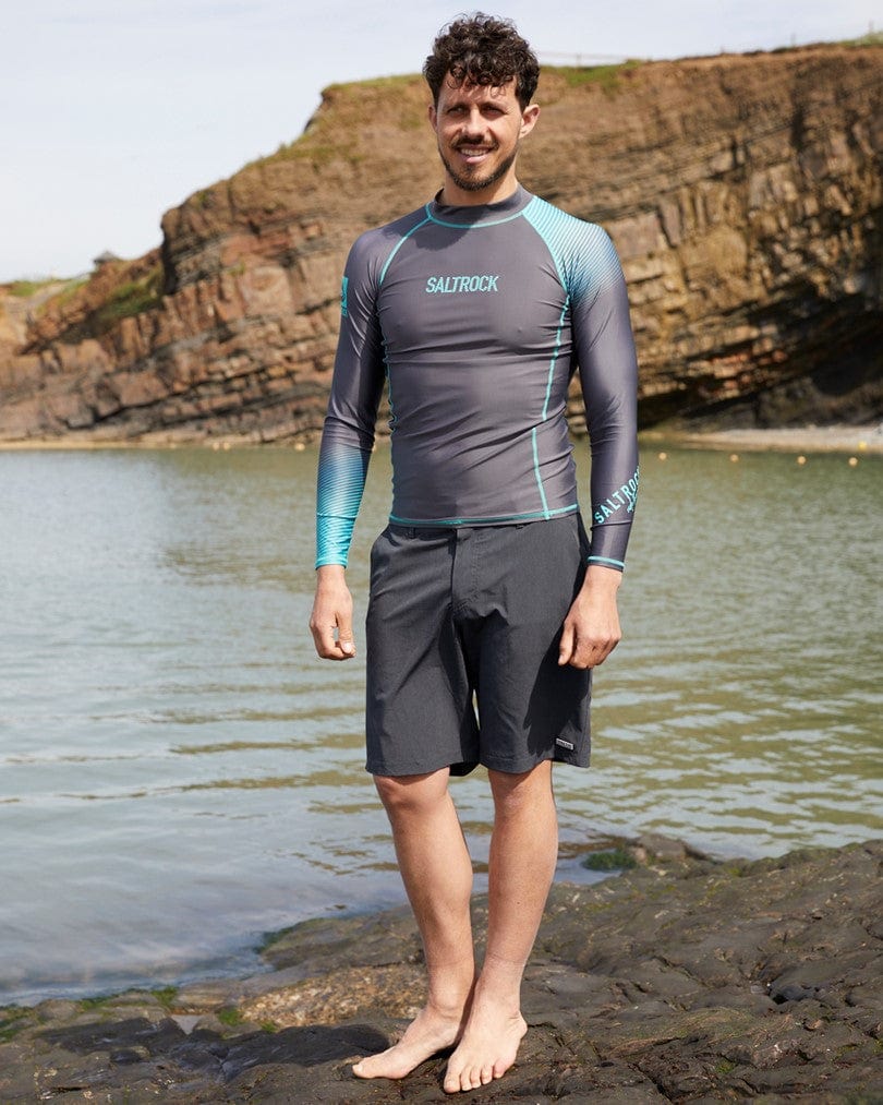 A man in a Saltrock DNA Wave - Mens Long Sleeve Rashvest - Grey/Turquoise and shorts standing on a rocky beach shoreline.
