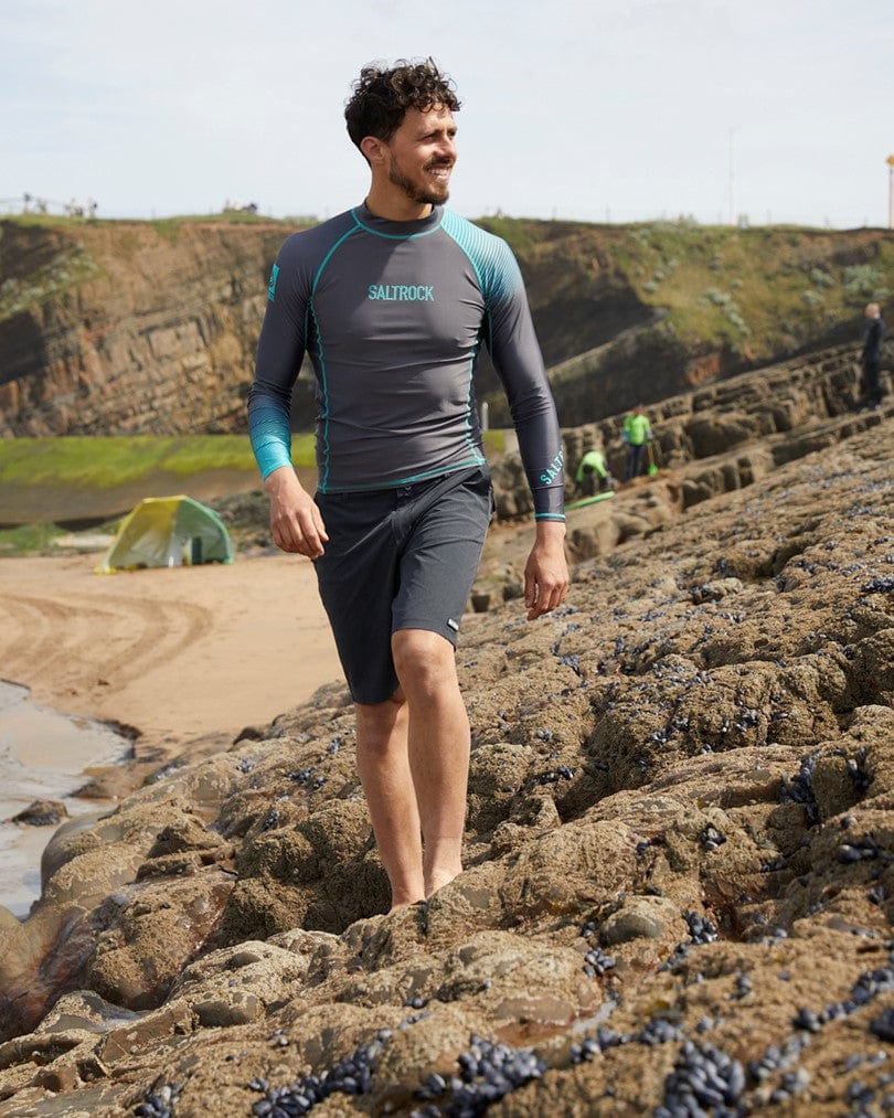 A man in a Saltrock DNA Wave Long Sleeve Rashvest in Grey/Turquoise walking on a rocky beach, with tents and cliffs in the background.