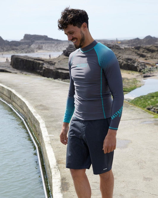 Man in a gray and teal Saltrock DNA Wave - Mens Long Sleeve Rashvest - Grey/Turquoise rash guard and black shorts standing by a seaside concrete walkway, smiling downward.