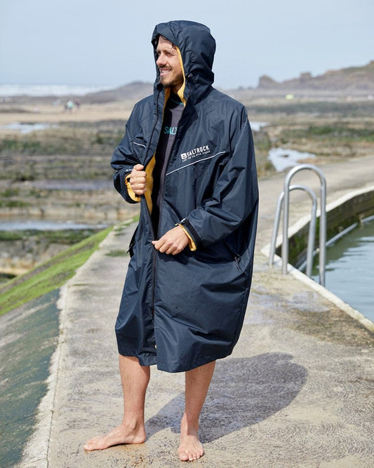 Man in a Saltrock Recycled Four Seasons Changing Robe - 3 in 1 - Black/Yellow standing barefoot by a seaside pool, smiling and holding a yellow mug.