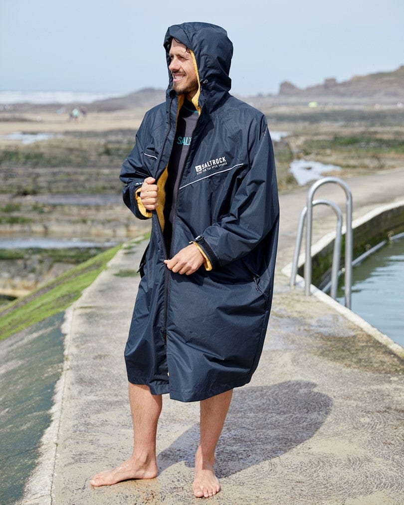 Man in a Saltrock black waterproof 3 in 1 Recycled Four Seasons Changing Robe standing barefoot by a seaside pool, holding a yellow mug, with a relaxed smile on his face.