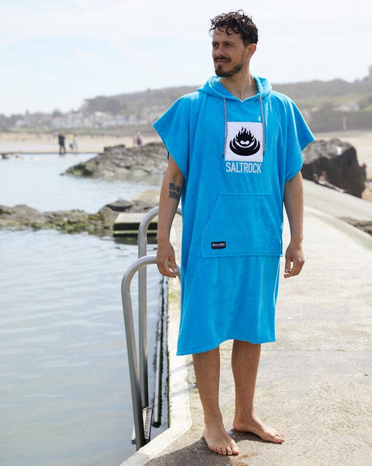 Man wearing a blue ultra absorbent Saltrock Corp Changing Towel standing by a seaside railing, with people and water in the background.