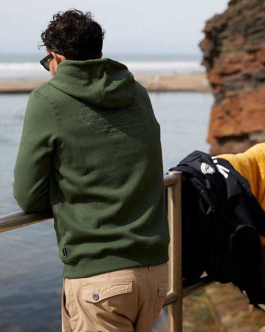 Man wearing a Saltrock Vantage Outline - Recycled Mens Zip Hoodie in Dark Green standing by a railing, facing a beach and cliff, with a black backpack hanging on the railing.