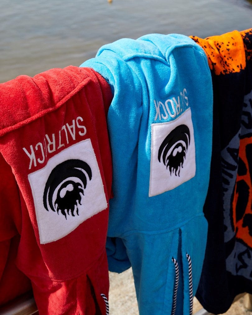 Colorful hoodies with Saltrock branding hanging to dry on a wooden railing by the water.