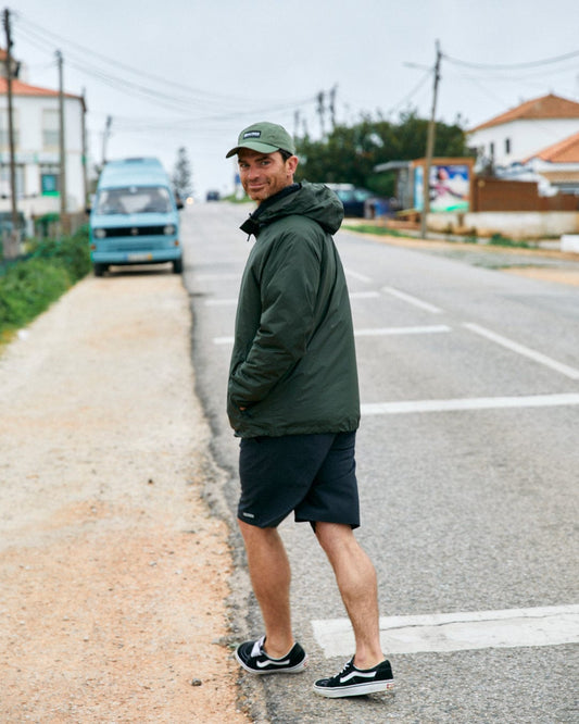 Man in a Saltrock Rainier - Mens Packable Waterproof Jacket in Green and black shorts smiling over his shoulder while standing on a suburban road.