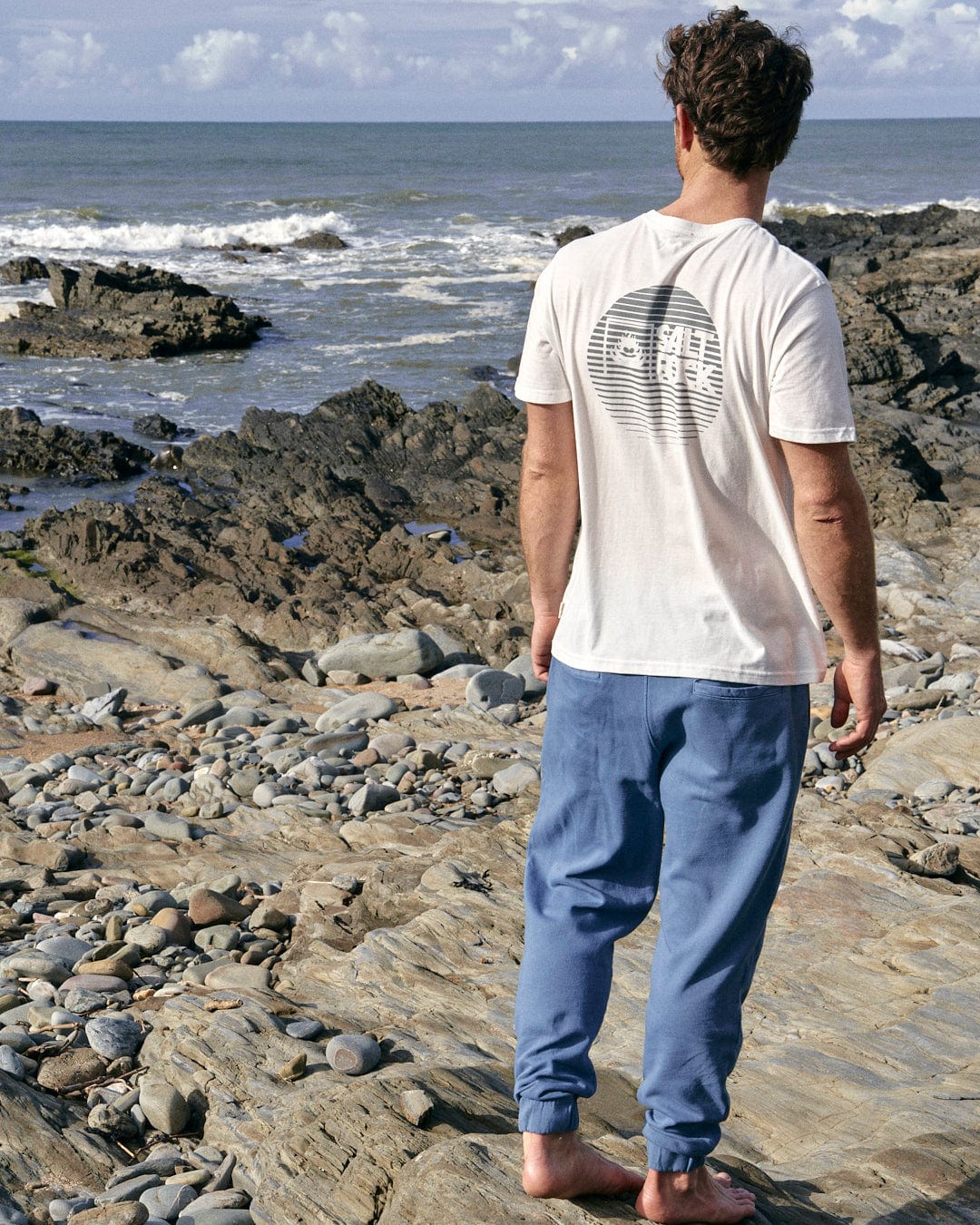A stylish man standing on a rocky beach with Saltrock's Ralfen - Mens Slimfit Sweatpants in Blue, holding a surfboard.