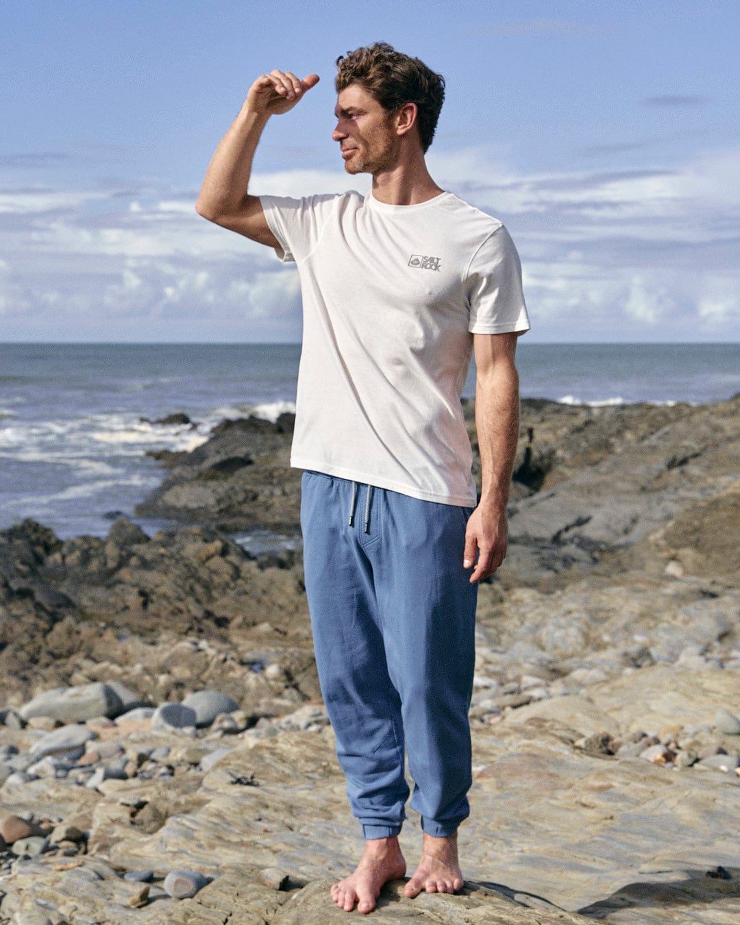 A stylish man standing on a comfortable beach wearing a white t-shirt and Saltrock's Ralfen - Mens Slimfit Sweatpants in Blue.