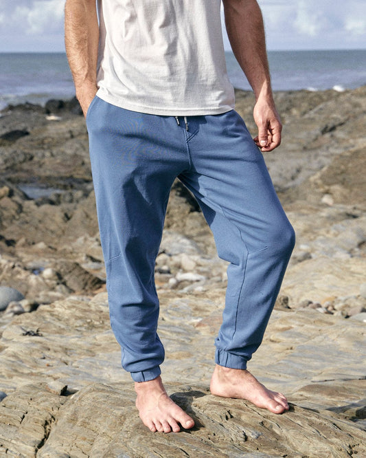 A man standing on rocks next to the ocean wearing Saltrock's Ralfen - Mens Slimfit Sweatpants in Blue.