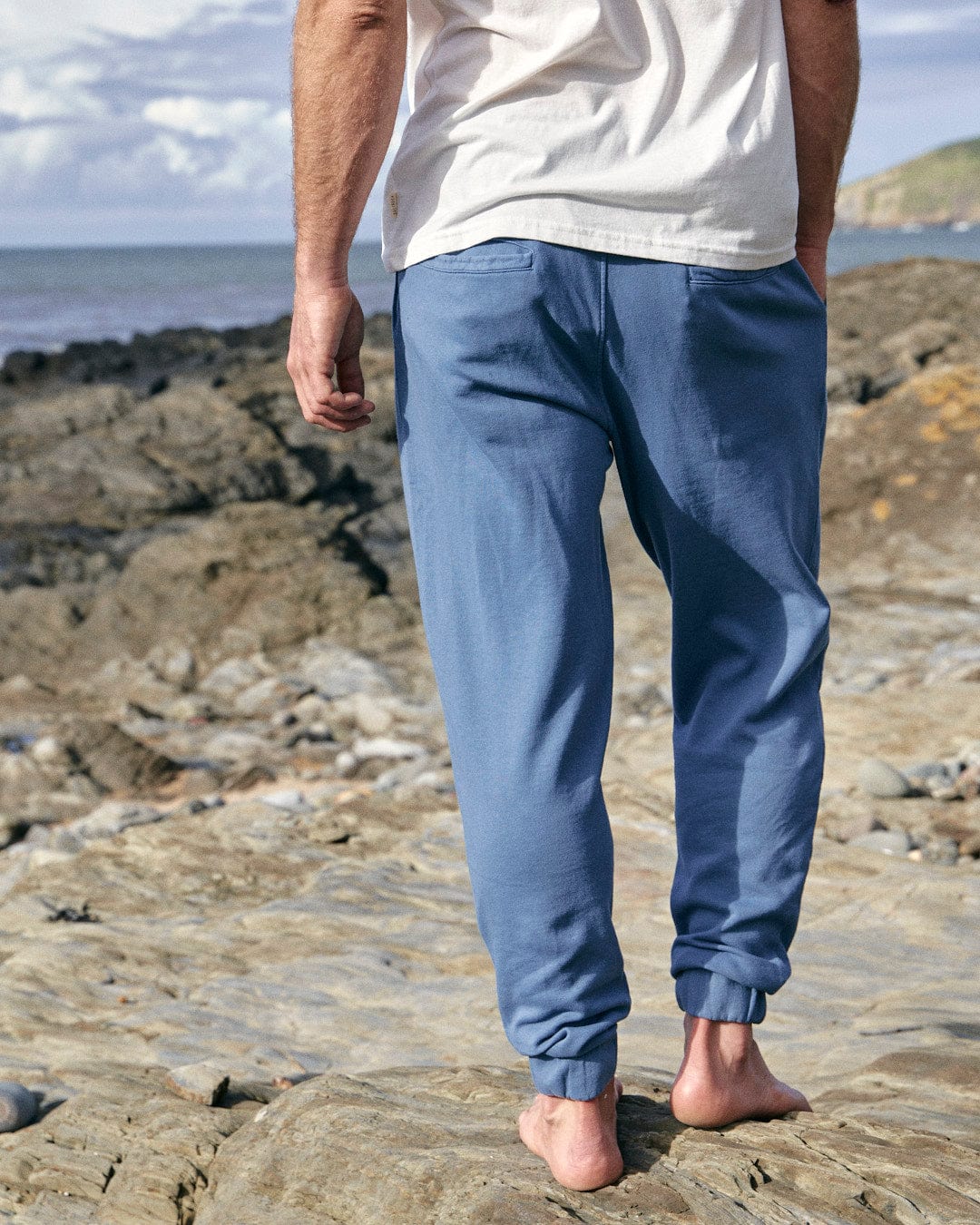 A man standing on a rocky beach wearing Saltrock's Ralfen - Mens Slimfit Sweatpants - Blue joggers.