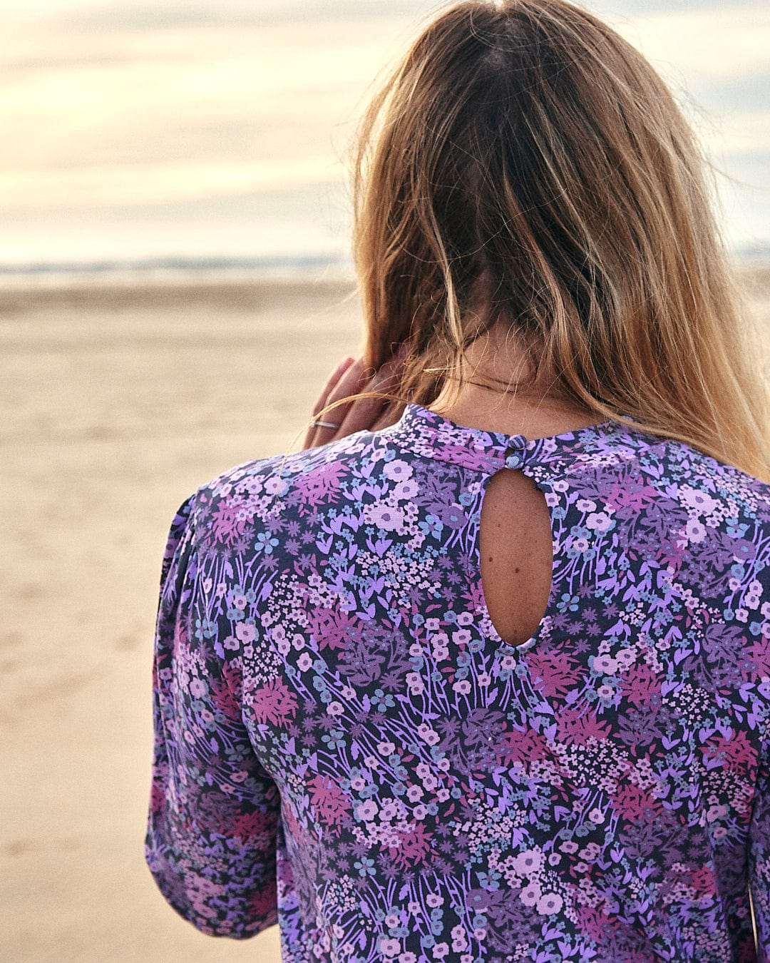 The back of a woman wearing a Saltrock Ramona - Womens Mock Jersey Dress - Purple with a purple floral print on the beach.