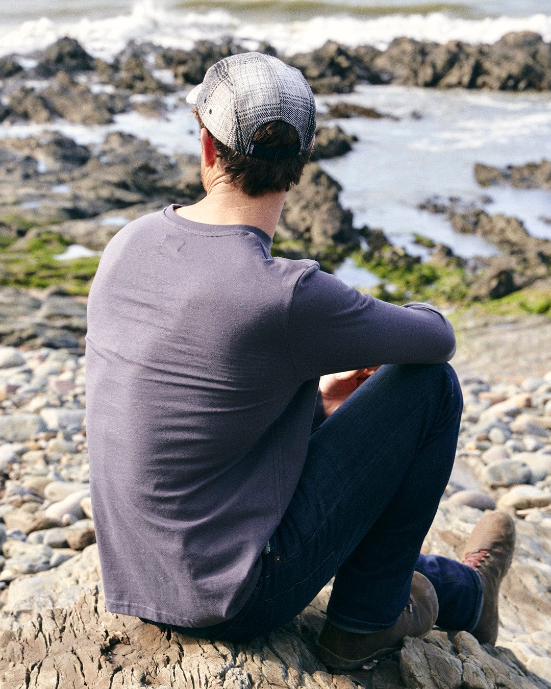 A man sitting on a rock, wearing a Saltrock Reflect - Mens Long Sleeve T-Shirt - Grey, looking at the ocean.