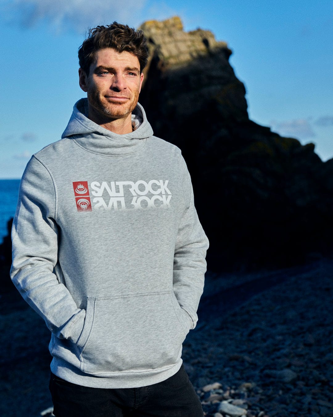 A man wearing a grey Reflect - Mens Pop Hoodie - Grey with Saltrock branding standing next to the ocean.