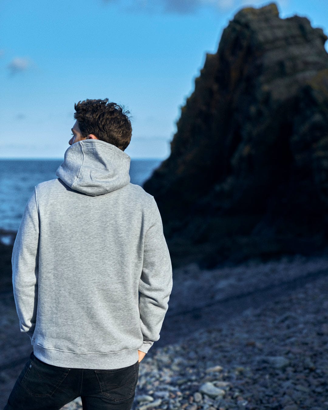 A man wearing a Reflect - Mens Pop Hoodie - Grey made by Saltrock, standing on a rocky beach showcasing Saltrock branding.
