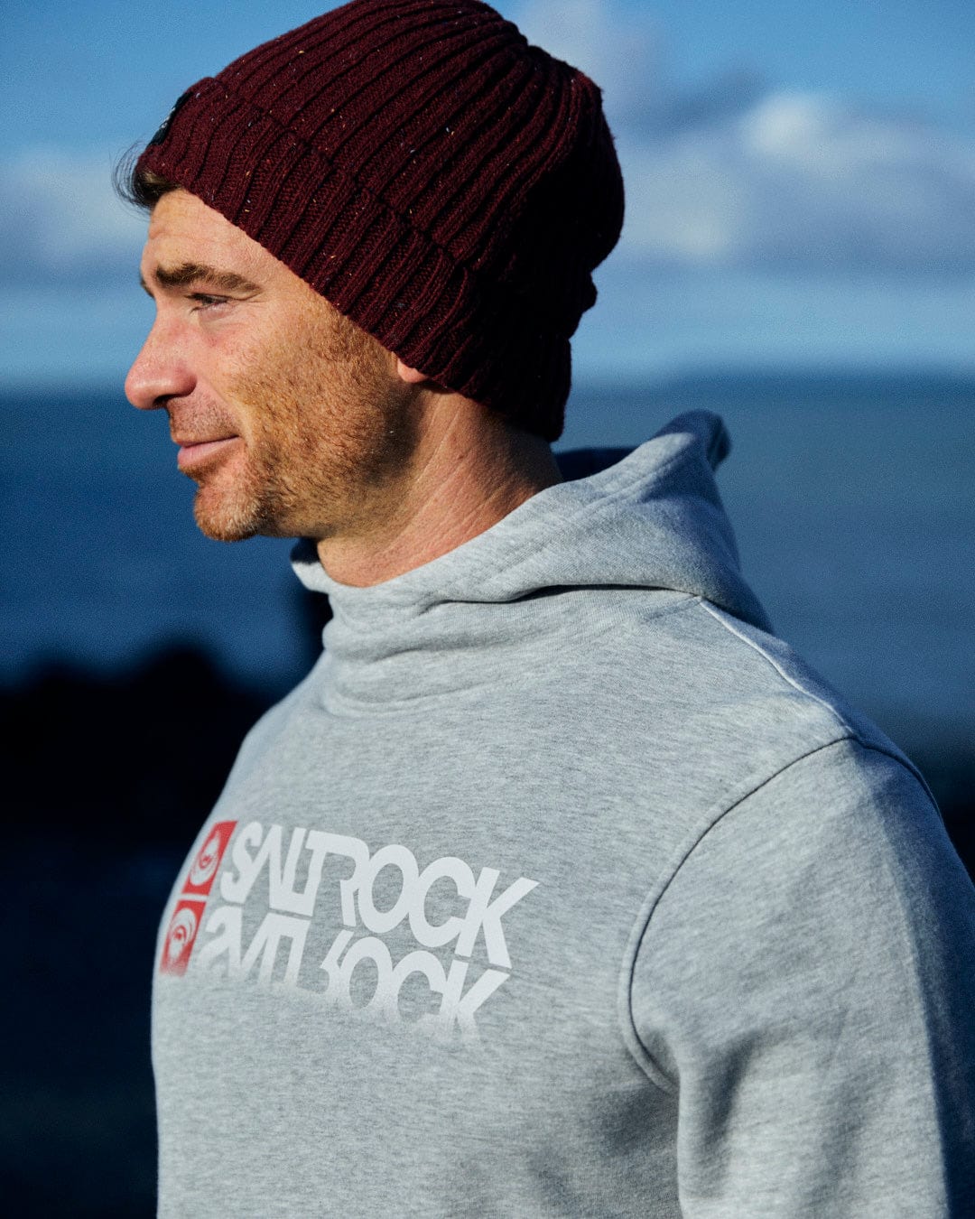 A man wearing a Reflect - Mens Pop Hoodie - Grey and a beanie in front of the ocean, showcasing Saltrock branding.