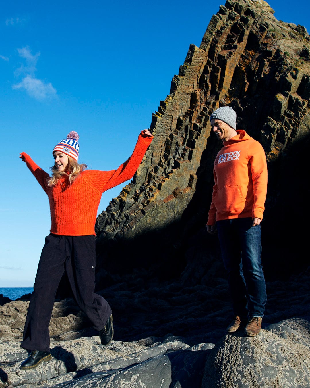 A man and woman, wearing a cotton blend Reflect - Mens Pop Hoodie - Orange with Saltrock branding, standing on a rocky beach with a frisbee.