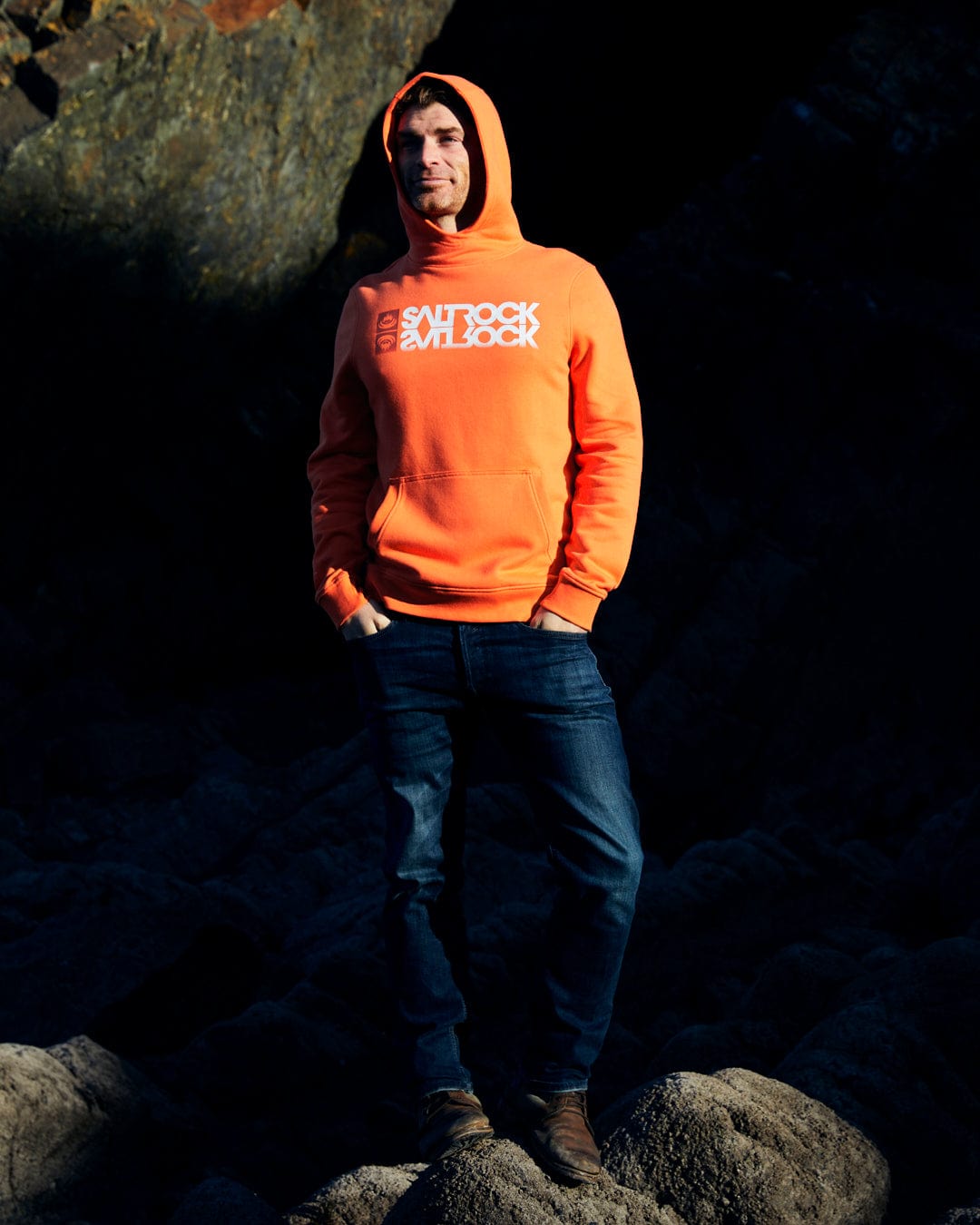A man in a Saltrock Reflect - Mens Pop Hoodie - Orange made from a comfortable cotton blend standing on rocks.