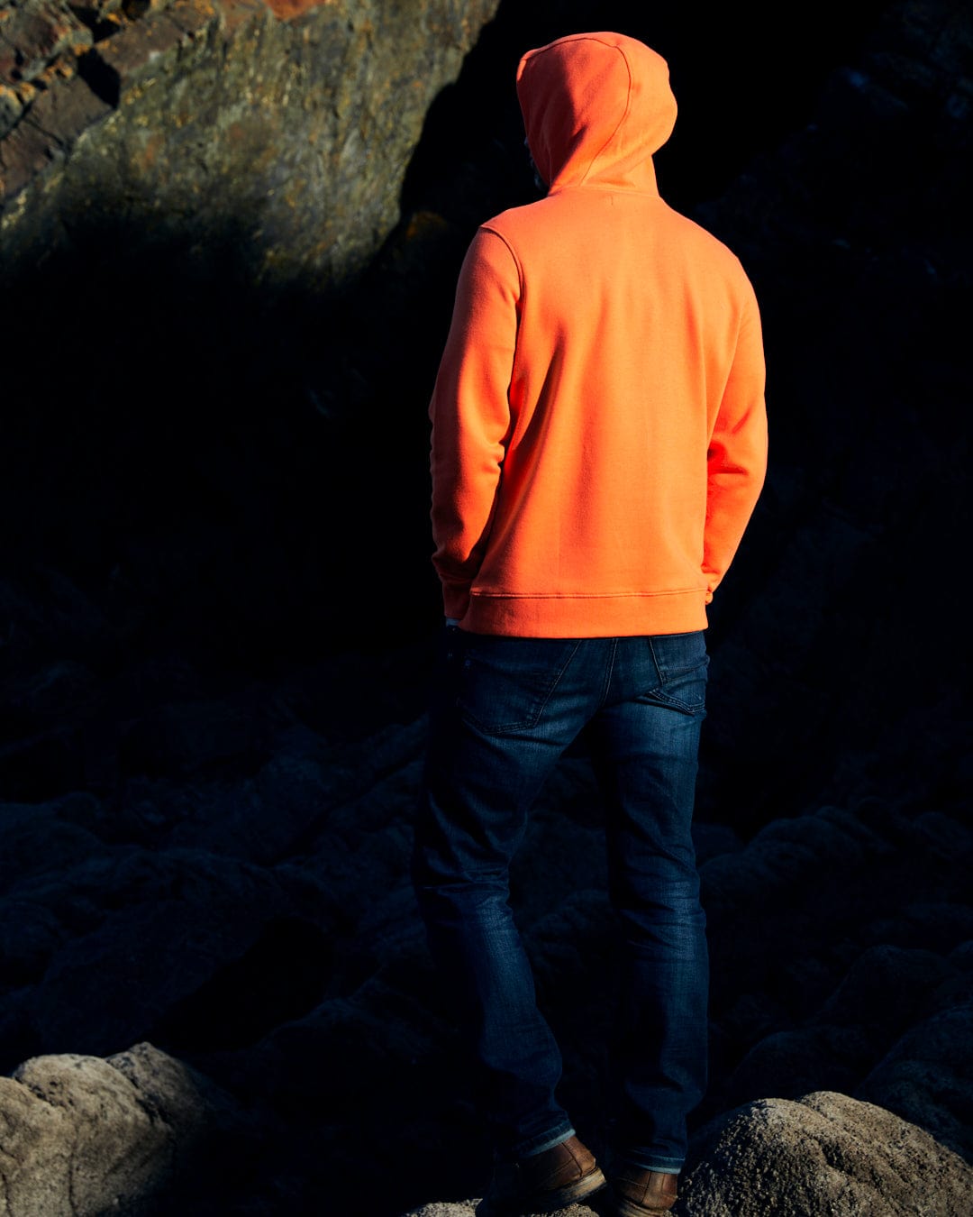 A man in an orange Reflect - Mens Pop Hoodie - Orange Saltrock branded hoodie standing on rocks.