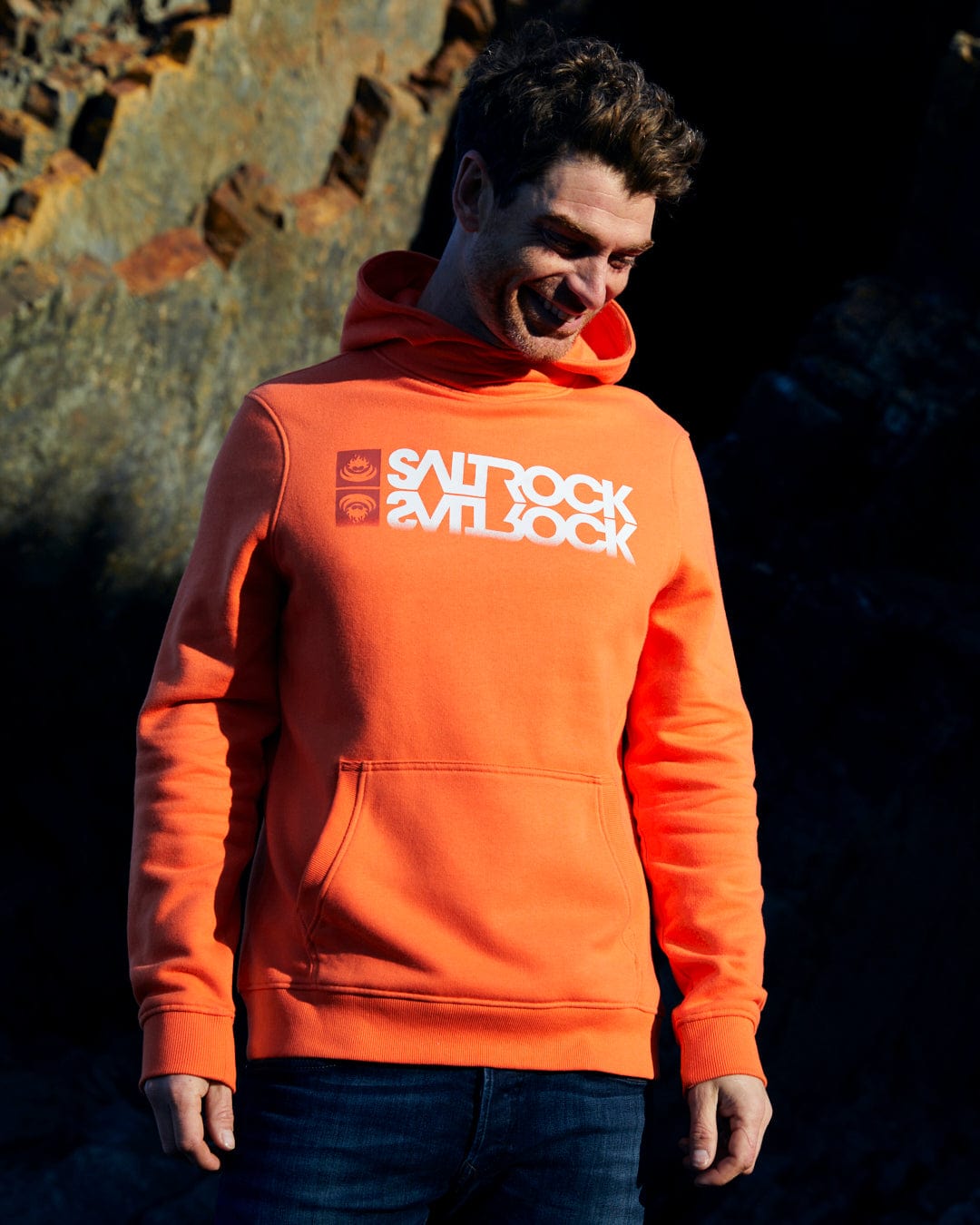 A man wearing a Saltrock Reflect - Mens Pop Hoodie - Orange, standing in front of rocks.