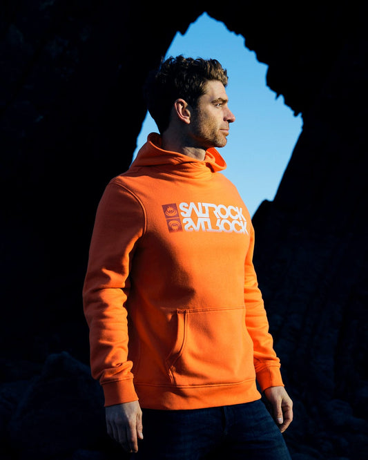A man wearing an orange Saltrock Reflect - Mens Pop Hoodie made from a comfortable cotton blend, standing in front of rocks.