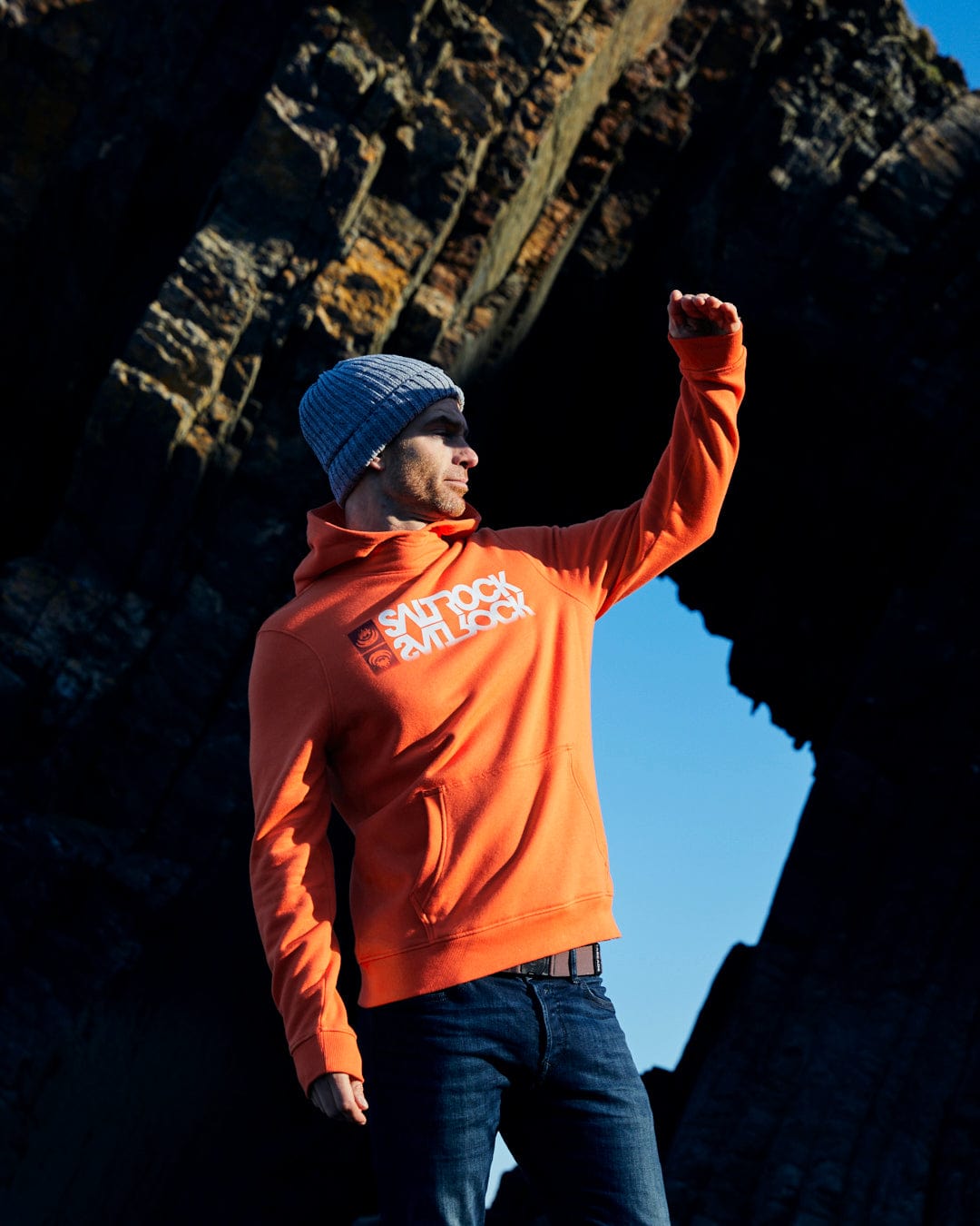 A man in a Reflect - Mens Pop Hoodie - Orange, showcasing the Saltrock branding with confidence amidst a scenic rocky backdrop.