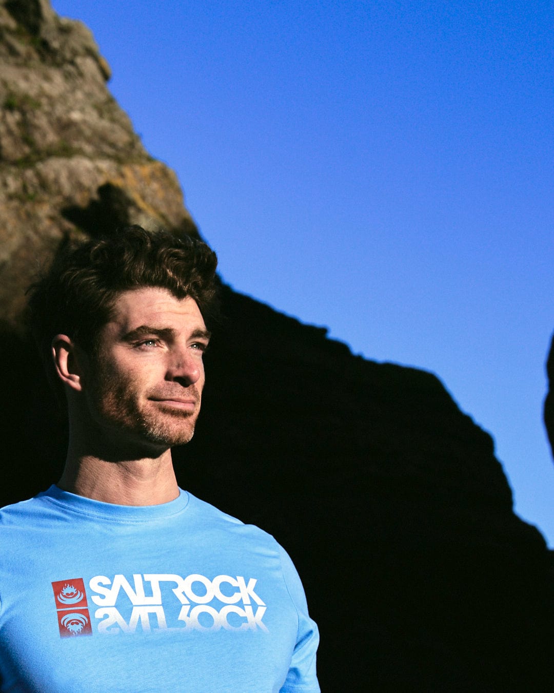 A man wearing a Saltrock Reflect - Mens T-Shirt - Blue standing in front of a rock.
