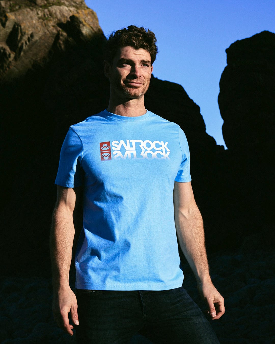 A man wearing a blue Reflect - Mens T-Shirt - Blue with Saltrock branding, standing in front of rocks.