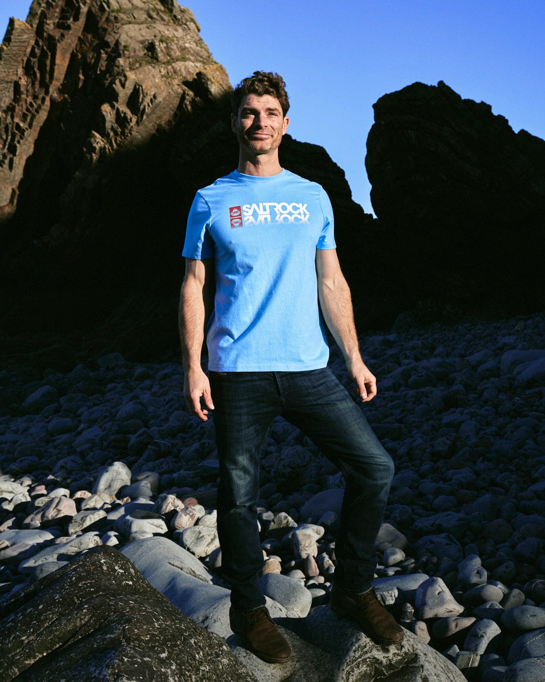 A man wearing a Saltrock Reflect - Mens T-Shirt - Blue standing on rocks.