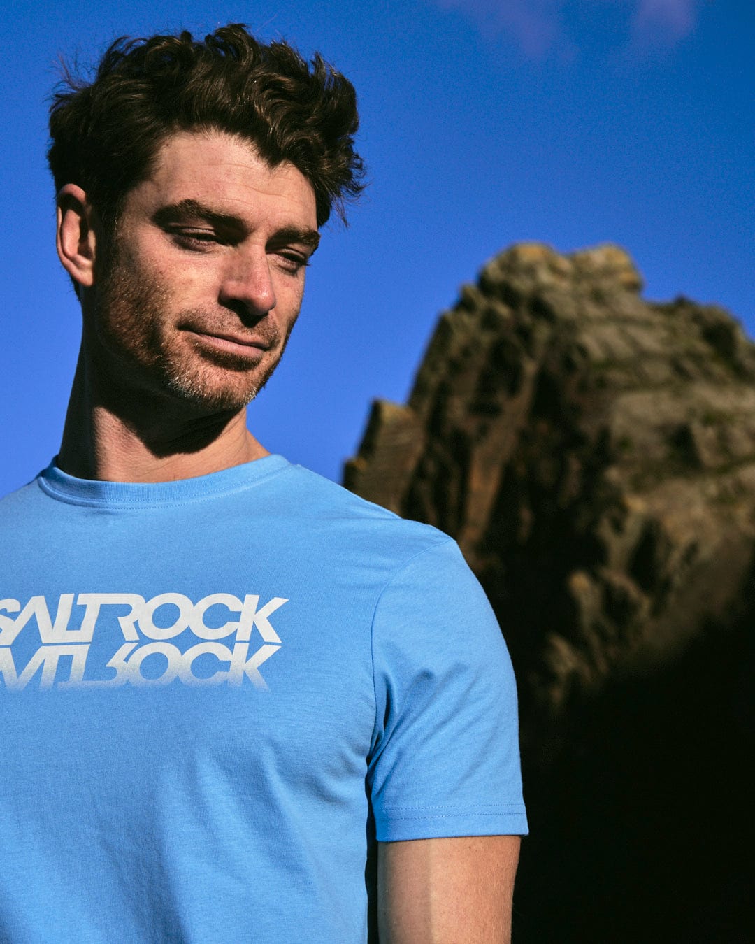 A man in a Saltrock Reflect Mens T-Shirt - Blue with a crew neck standing in front of a rock.