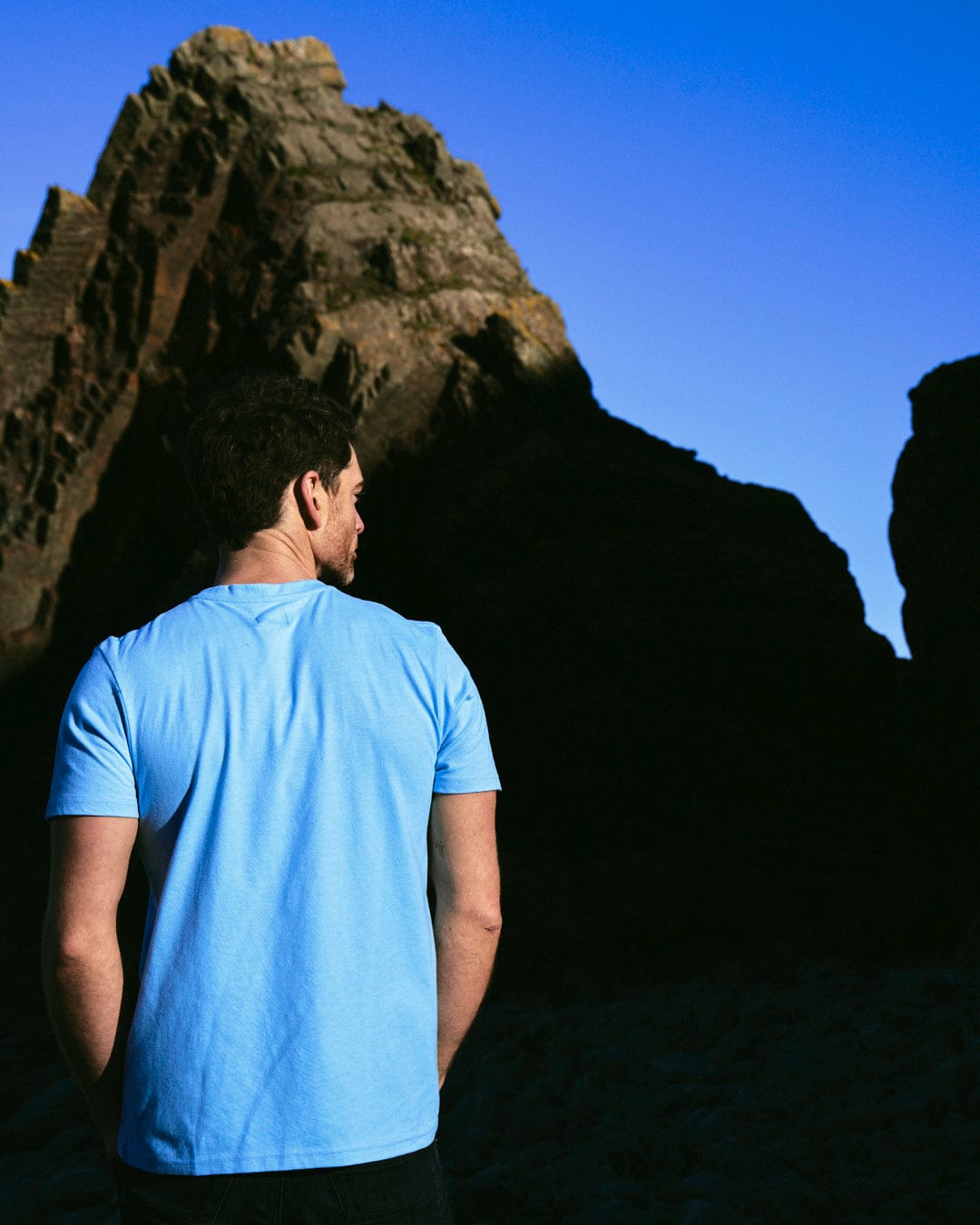 A man wearing a blue Saltrock Reflect Mens T-Shirt, standing in front of a rock.