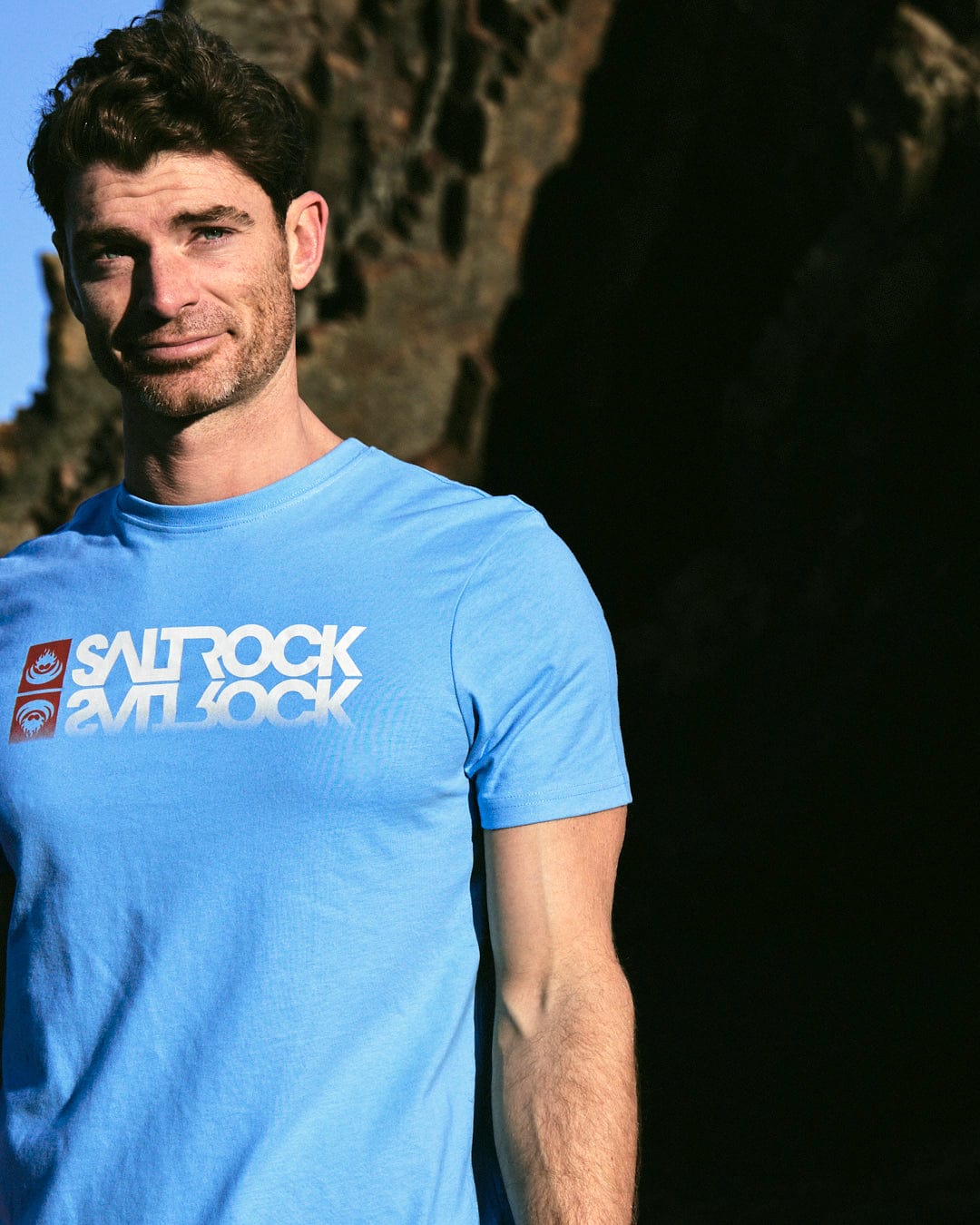 A man wearing a Saltrock Reflect Mens T-Shirt in Blue standing in front of a rock.