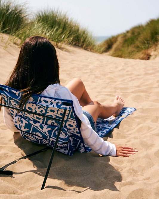 A woman sitting on a beach chair in the sand, enjoying the comfort of a Saltrock Relax - Beach Mat - Blue with its fold away design.