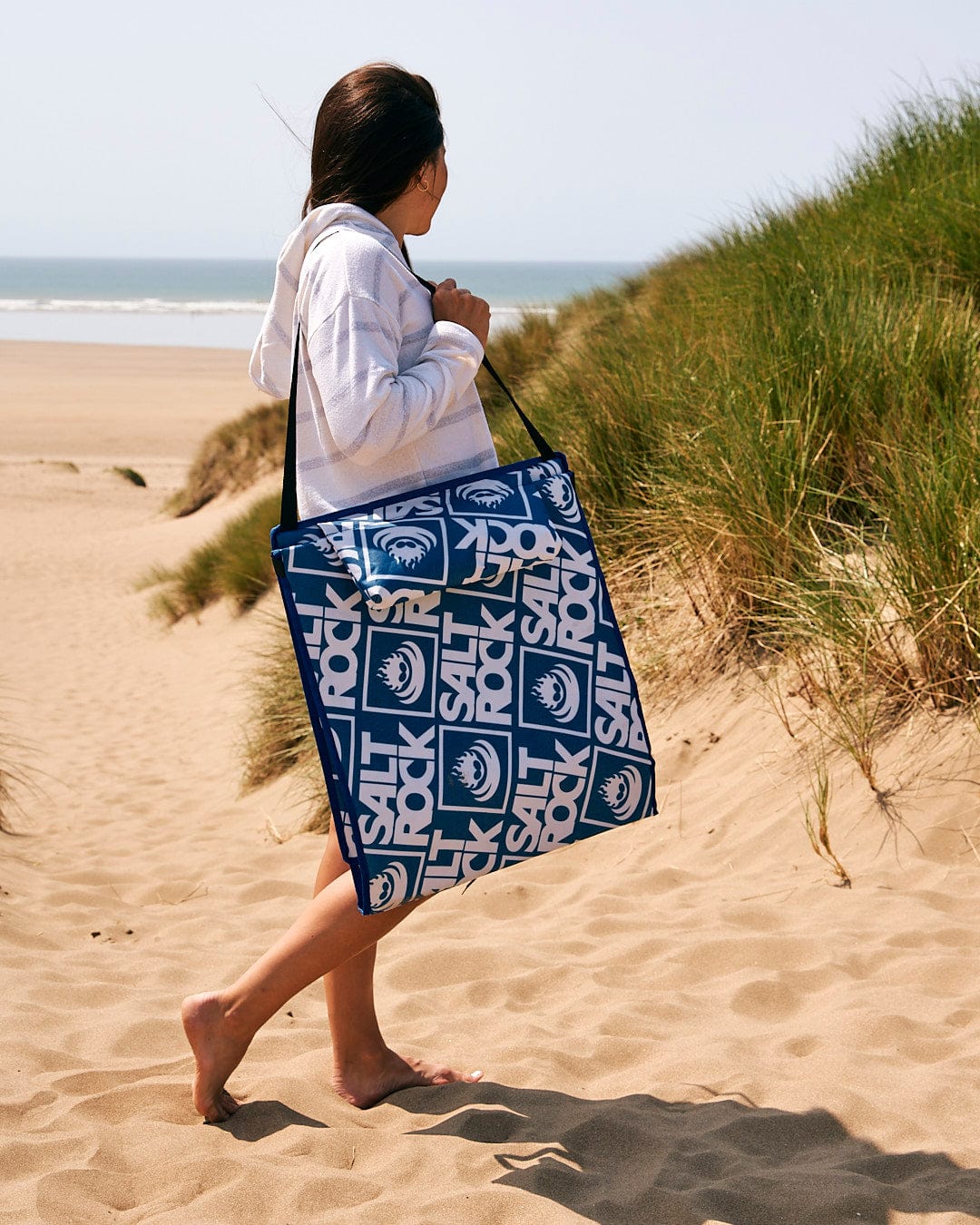 A woman carrying a Saltrock Relax - Beach Mat - Blue with a fold away design on the sand.