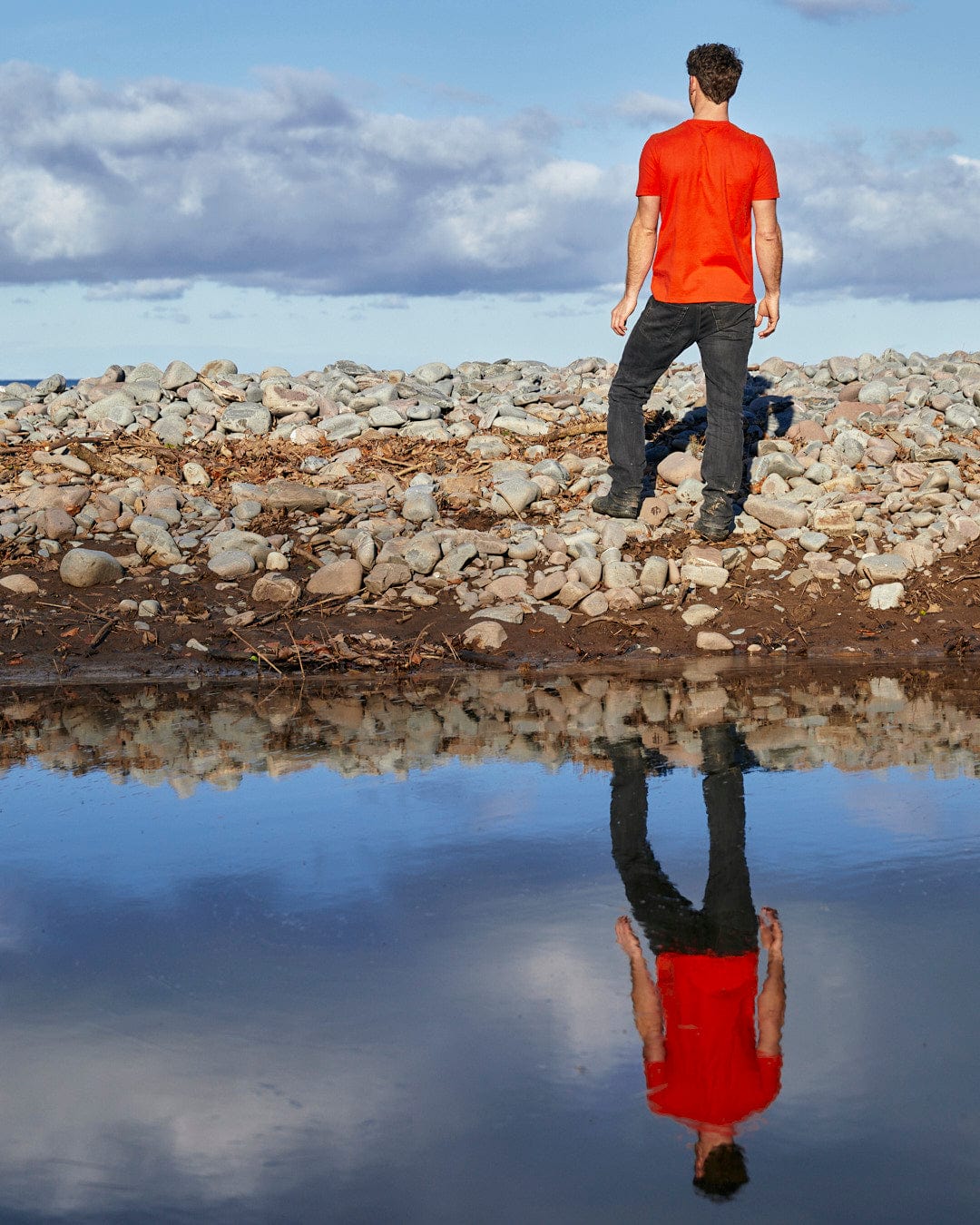 A man, sporting a Saltrock Retro Carve - Mens Short Sleeve T-Shirt - Red design on his shirt, is standing on a rocky shore looking at his reflection in the water.