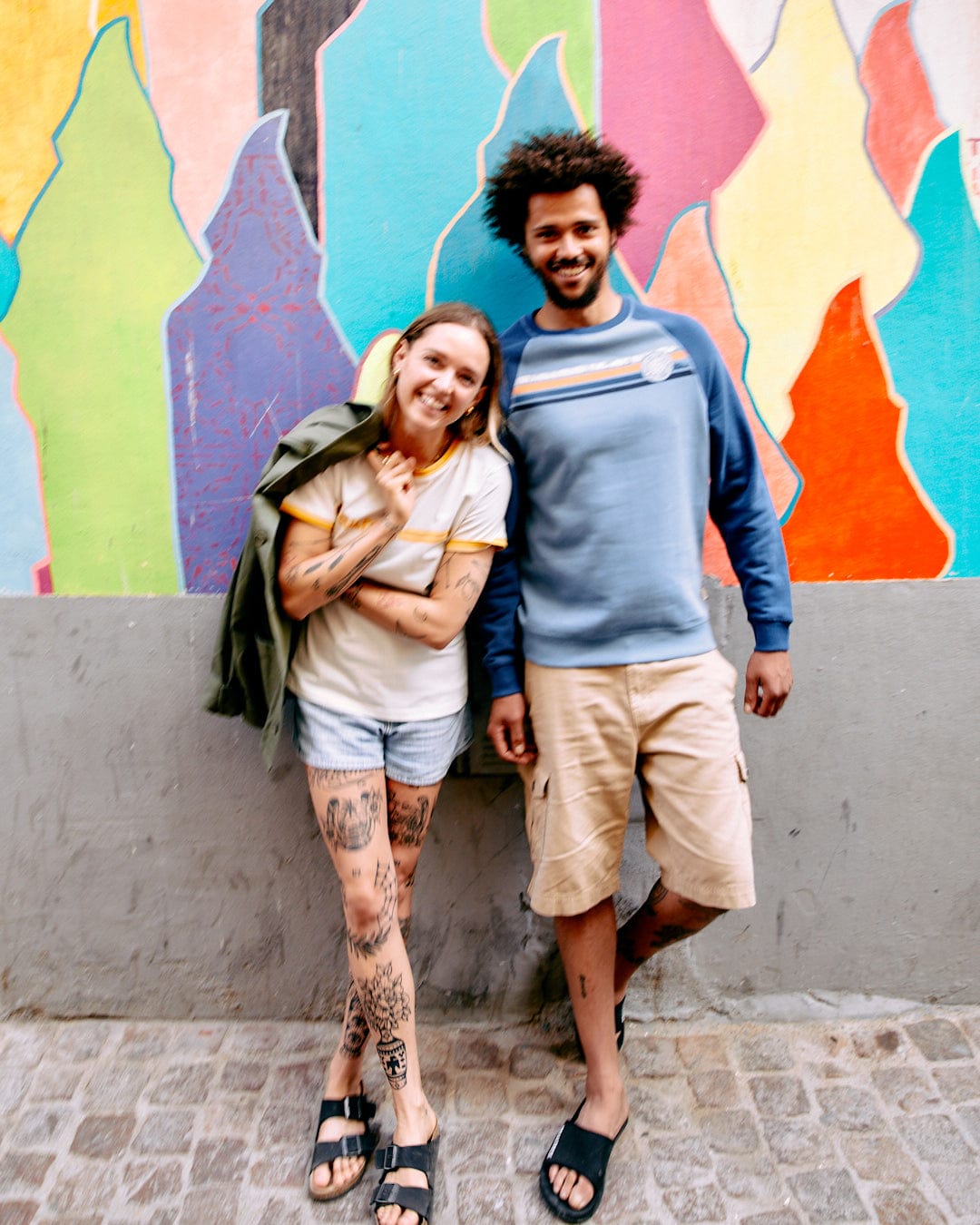 A smiling young couple standing in front of a colorful mural, the man with afro hair and the woman displaying tattooed legs and a Saltrock Retro Ribbon - Womens Shorts Sleeve T-Shirt in White.