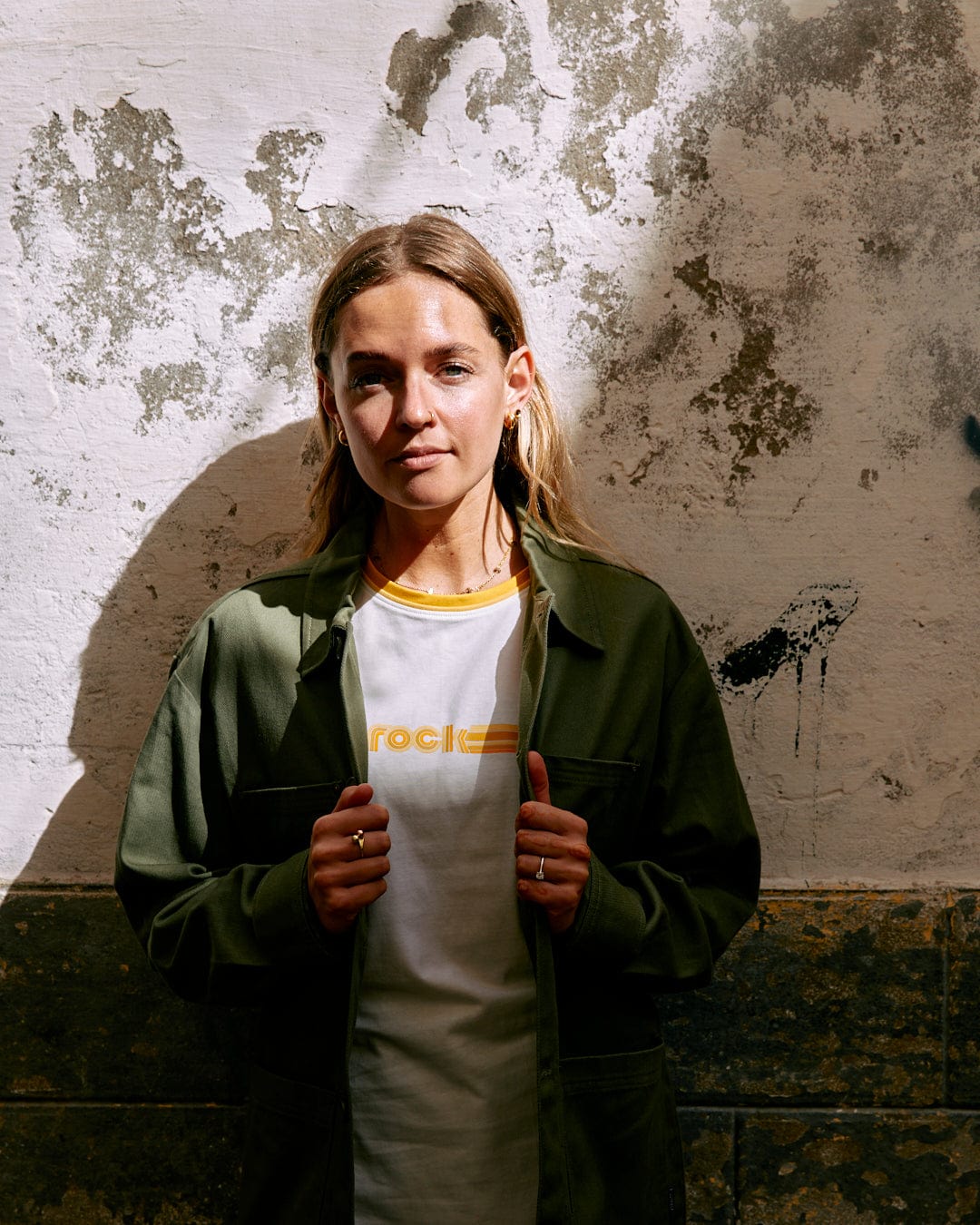 Young woman in a green jacket and Retro Ribbon - Womens Shorts Sleeve T-Shirt - White by Saltrock standing against a textured wall, sunlight casting shadows on her face.