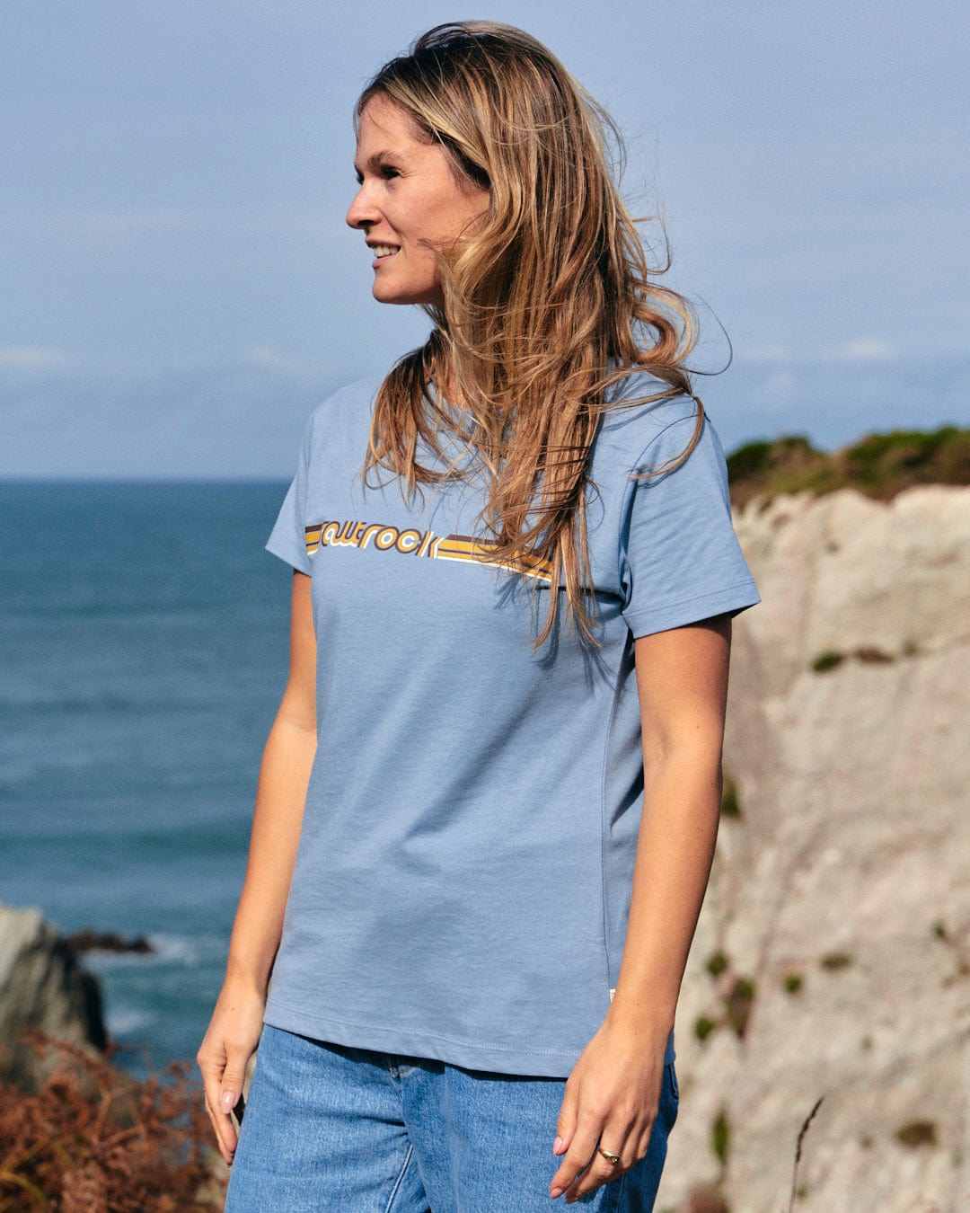 A woman wearing a blue Saltrock Retro Ribbon - Womens Short Sleeve T-Shirt - Light Blue standing next to the ocean.