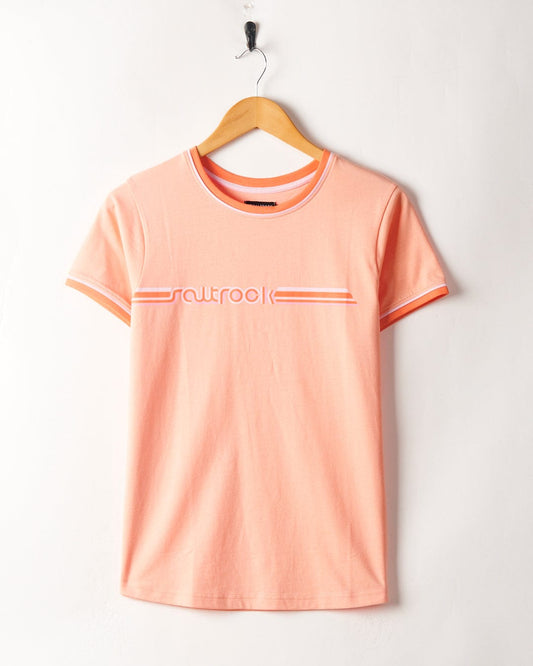 A Retro Ribbon - Womens Shorts Sleeve T-Shirt in peach color with "outlook" printed across the chest, featuring retro stripes, hanging on a wooden hanger on a white wall by Saltrock.