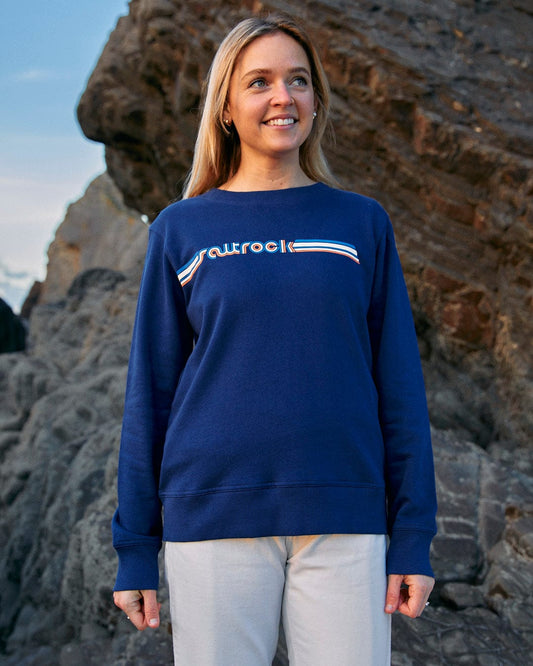 A woman wearing a Retro Ribbon - Womens Sweat - Blue sweatshirt branded as Saltrock, standing on rocks.