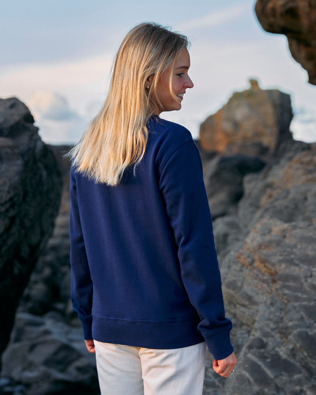A woman wearing a Retro Ribbon - Womens Sweat - Blue sweatshirt standing on Saltrock branded rocks.