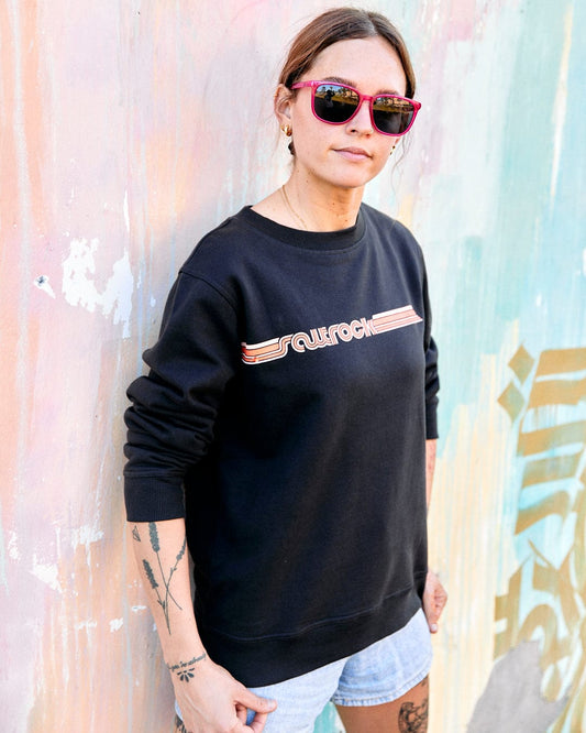 A woman in sunglasses and a Saltrock Retro Ribbon Tape - Womens Sweatshirt in Dark Grey stands against a colorful graffitied wall.