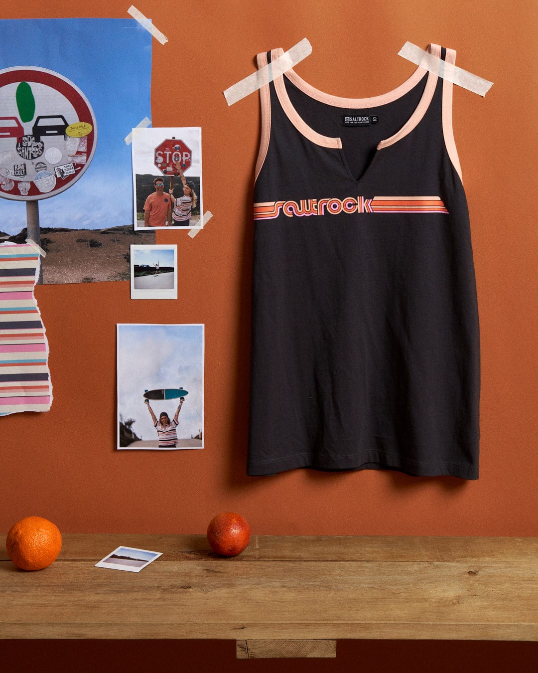 Dark grey tank top with "surfrock" logo and retro ribbon stripe, hanging on a wooden peg against an orange wall, decorated with various photographs and two oranges resting on a shelf below.