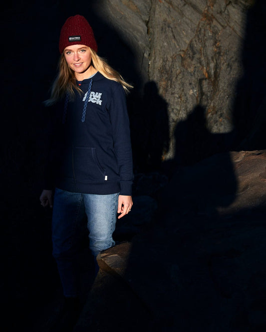 A woman in a Saltrock Retro Wave Emb - Womens Zip Hoodie - Blue with front pockets, standing on a rock.