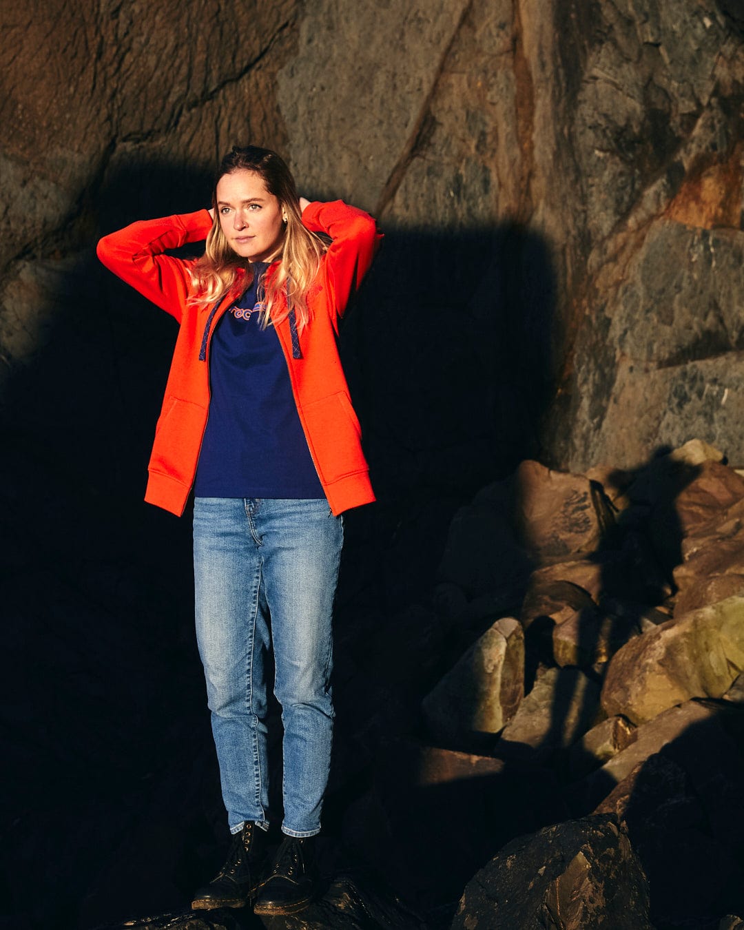 A woman in a Retro Wave Embroidered - Womens Zip Hoodie - Red, featuring subtle Saltrock branding and contrast blue hood lining, standing on rocks.