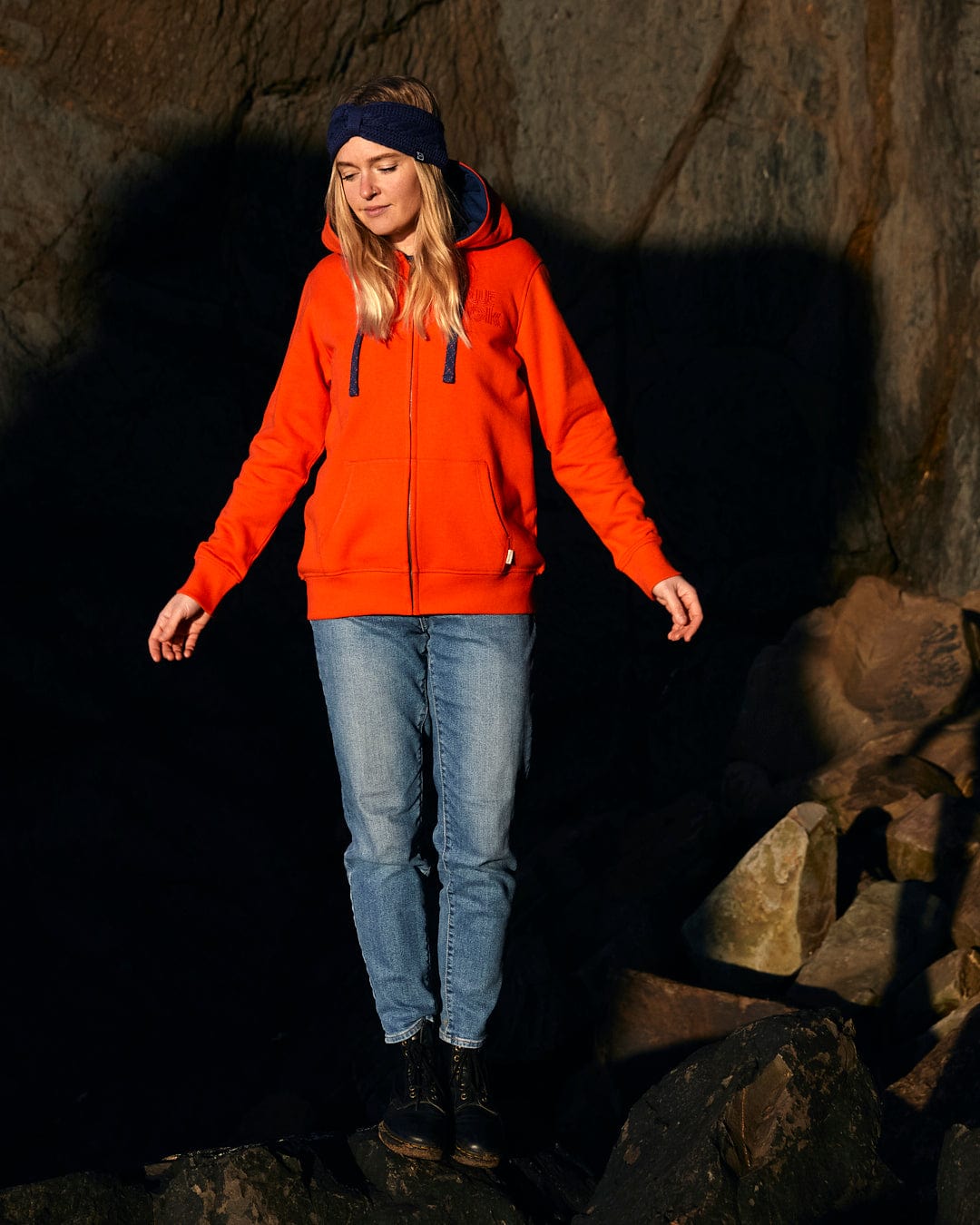 A woman in a Retro Wave Embroidered - Womens Zip Hoodie - Red Saltrock standing on rocks, featuring subtle Saltrock branding and contrast blue hood lining.