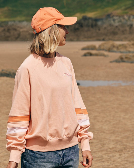 A person wearing the Saltrock "Retro Ribbon Block - Womens Sweatshirt - Peach" and a matching cap stands on a sandy beach with rocky hills in the background, adding a touch of coastal style to their cotton outfit.