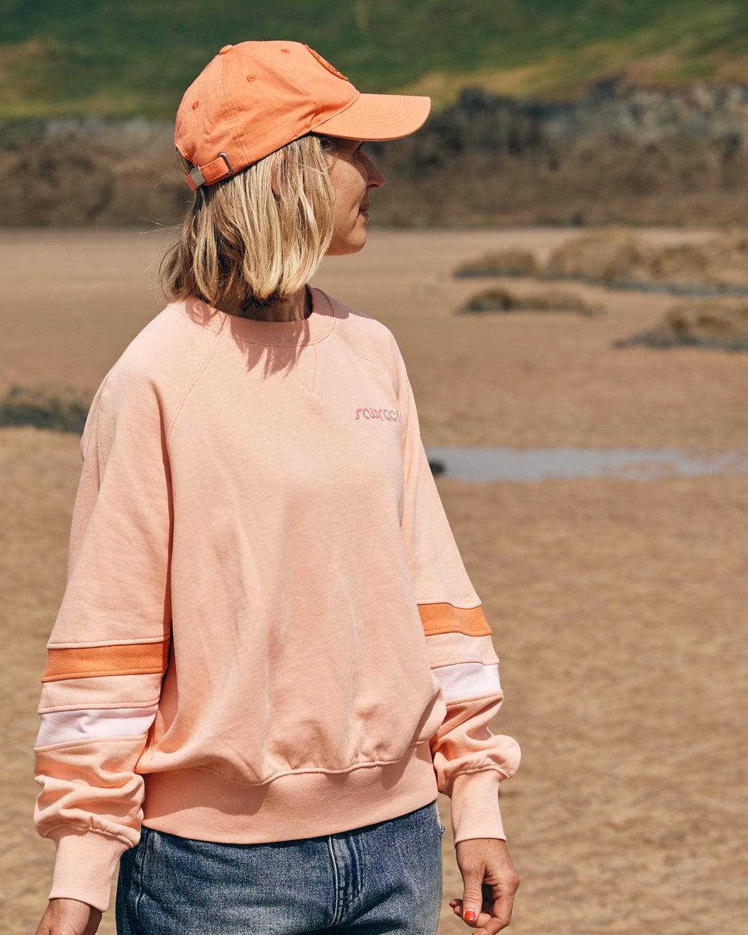 A person wearing the Saltrock "Retro Ribbon Block - Womens Sweatshirt - Peach" and a matching cap stands on a sandy beach with rocky hills in the background, adding a touch of coastal style to their cotton outfit.