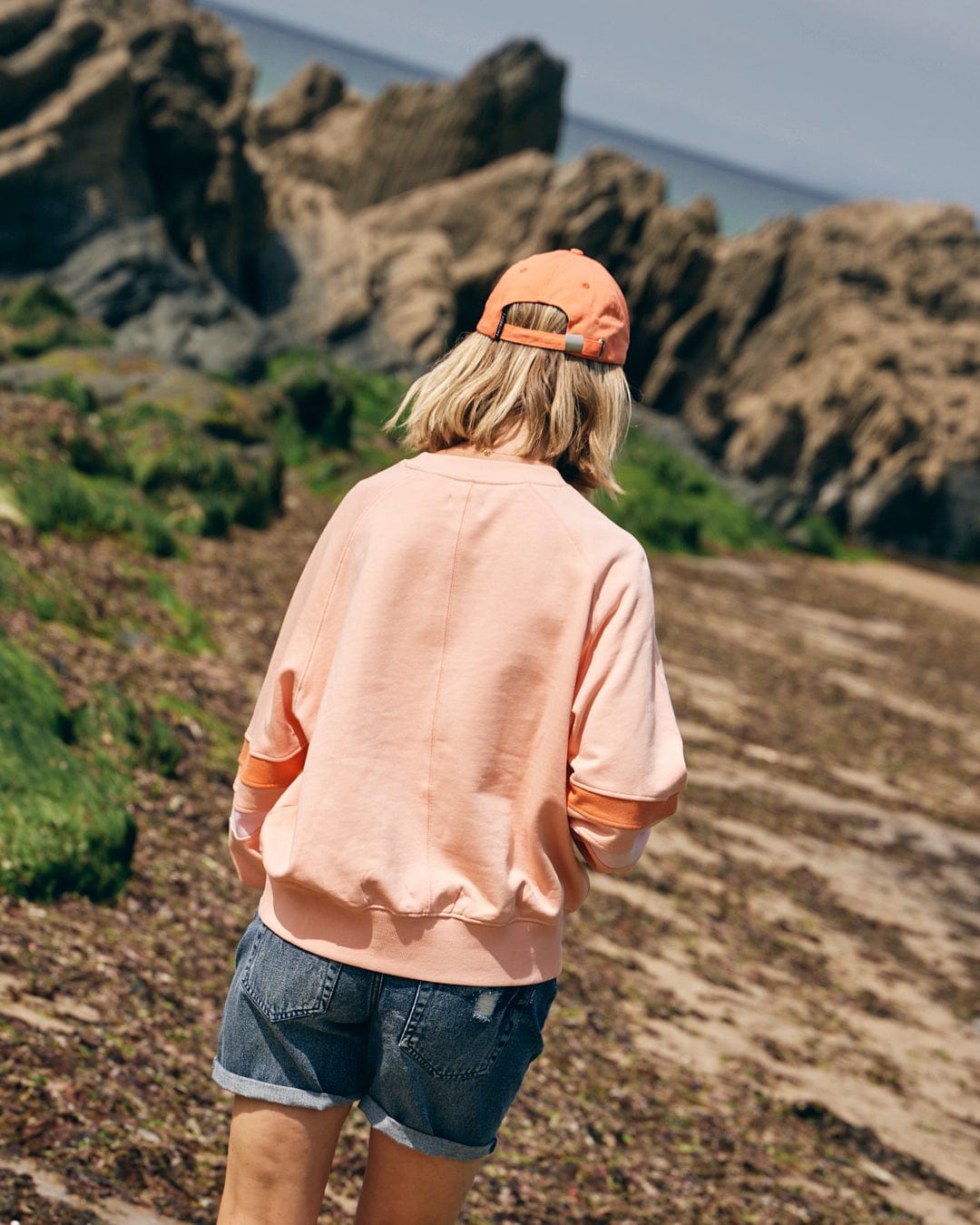 A person with blonde hair, wearing an orange cap, a Saltrock Retro Ribbon Block Women's Sweatshirt in peach, and denim shorts walks along a rocky shoreline.
